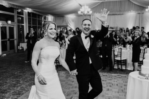 Bride and groom in a tented venue, smiling and waving amid applauding guests.