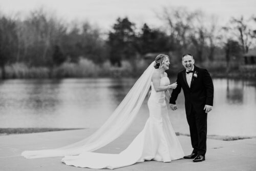 Bride and groom laughing by a lake, with the bride wearing a long veil. Black and white photo.