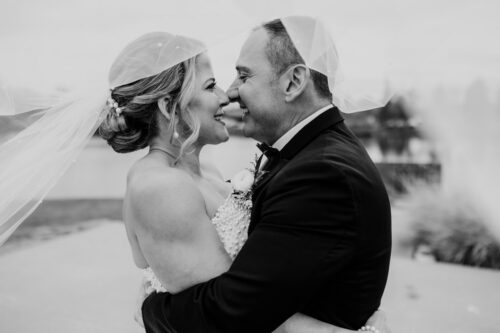 Black and white photo of bride and groom smiling under a veil, embracing each other outdoors before their Lincolnshire Marriott wedding ceremony