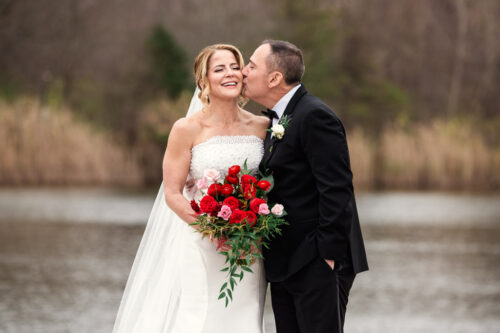Bride smiling as groom kisses her cheek by the lake at Lincolnshire Marriott Resort; she holds a bouquet of red and pink roses.