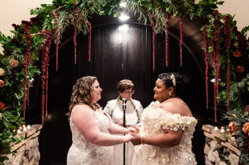Couple holds hands under a floral arch during their Osteria Via Stato wedding ceremony, with an officiant standing behind them