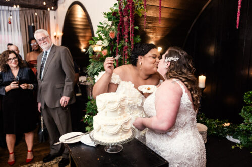 Brides in wedding dresses kiss after cutting their wedding cake, with onlookers smiling in the background