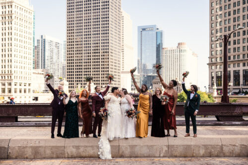 Wedding party cheers while brides kiss on a city street, holding bouquets and smiling against a Chicago skyline backdrop