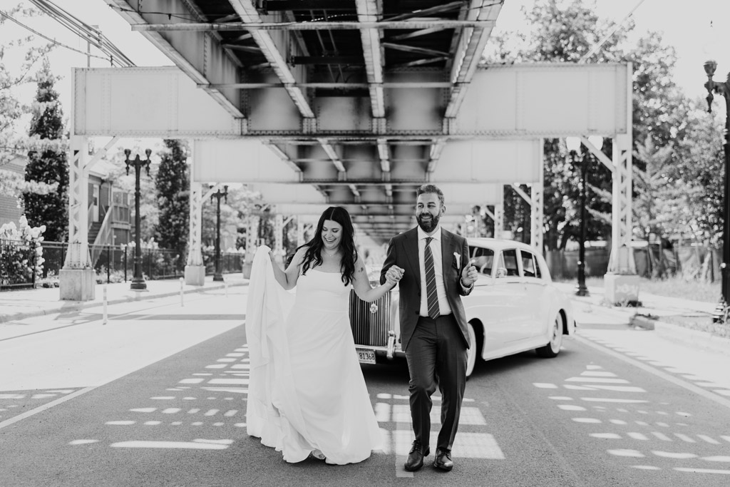 B;ack and white photo of bride and groom walking under the Lake Street El tracks beside a vintage car near Ovation Chicago