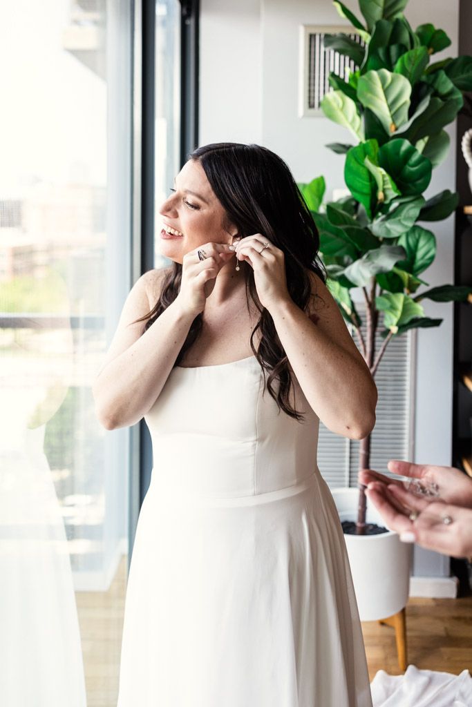Bride smiles as she puts on earrings near a window, wearing a white dress before her Ovation Chicago wedding celebration