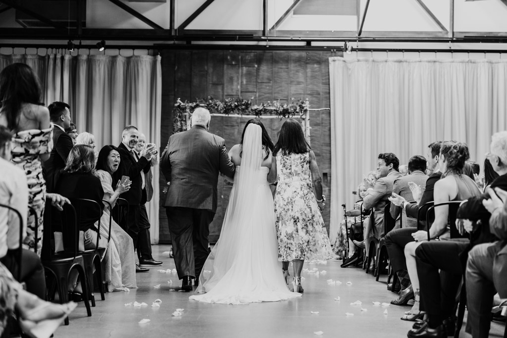 Black adn white photo of bride walking down the aisle with her parents at Ovation Chicago wedding ceremony venue, guests seated on either side