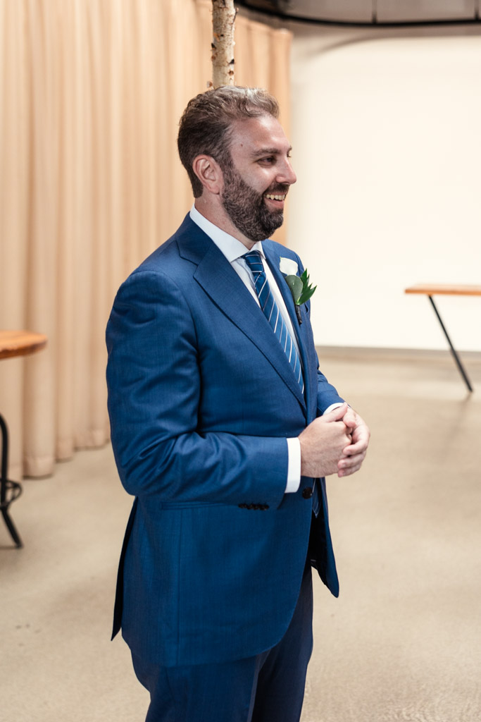 Groom in a blue suit smiling and standing indoors, hands clasped together, watching bride walk down the aisle during their Ovation Chicago wedding ceremony