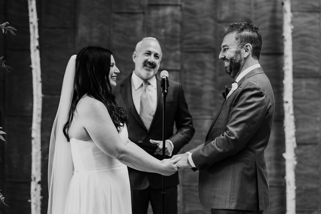 Black and white photo of bride and groom holding hands and smiling during their Ovation Chicago wedding ceremony with officiant visible in the background