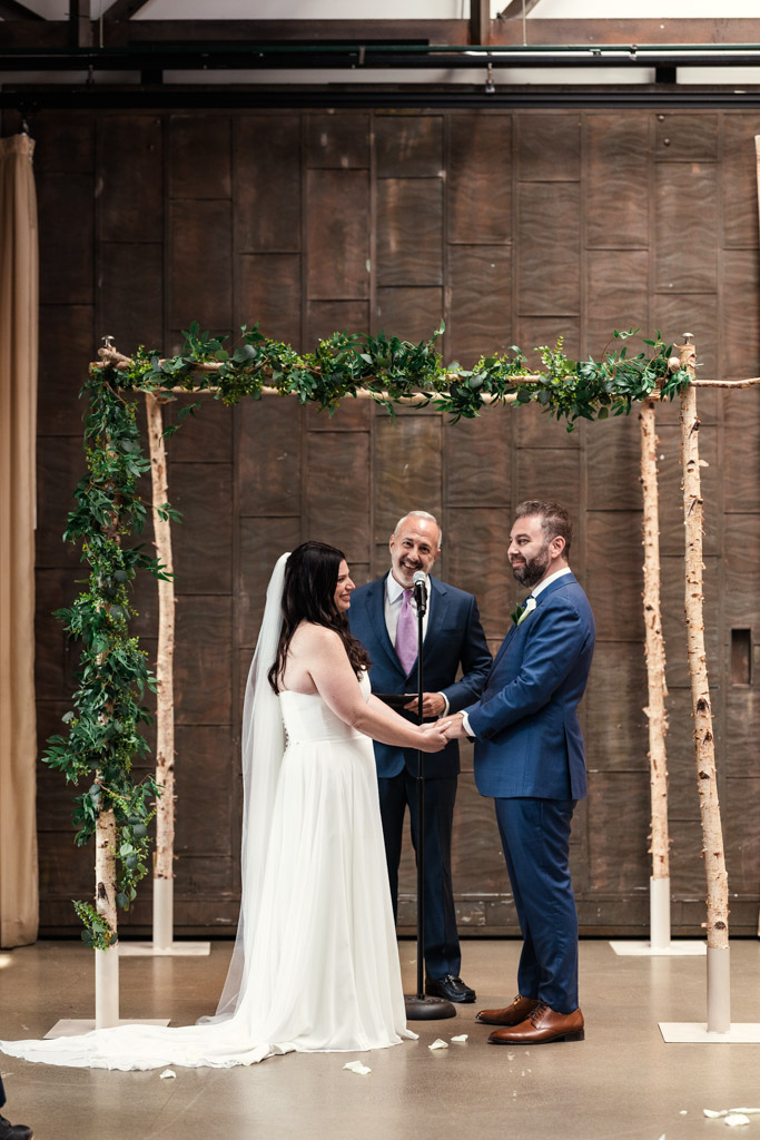 Bride and groom smile and hold hands under the chuppah while the officiant speaks during their Ovation Chicago wedding ceremony