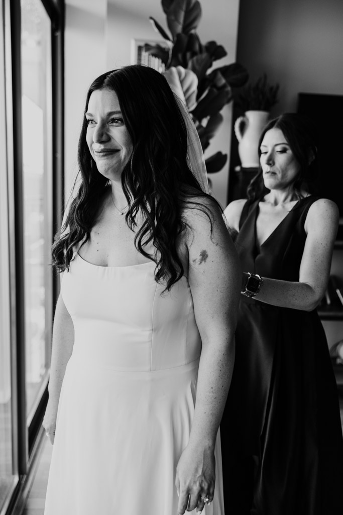 Black and white photo of bride smiling, while Maid of Honor adjusts her dress as they get ready at the Westin River North in downtown Chicago