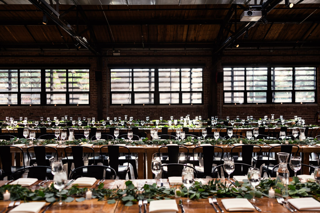 Wedding reception space at Ovation Chicago featuring long wooden tables and greenery centerpieces illuminated by natural light pouring through large windows