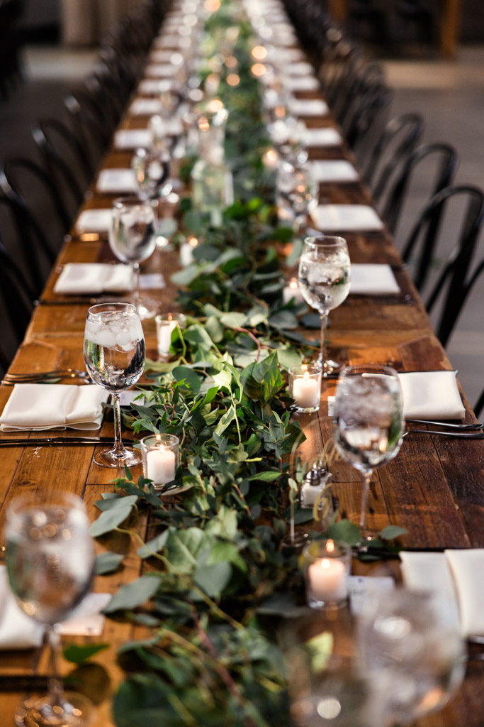 A long wooden guest table set with glassware, candles, and lush greenery for Ovation Chicago wedding reception