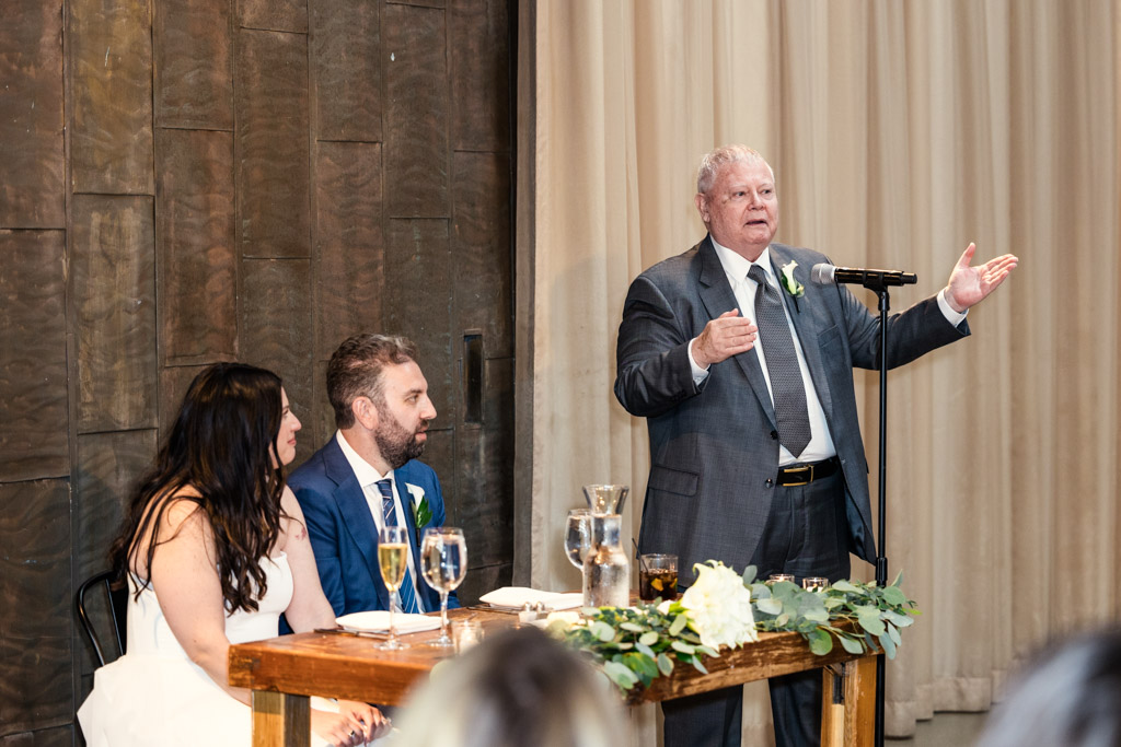 Gueat gives speech at Ovation Chicago wedding reception while bride and groom listen