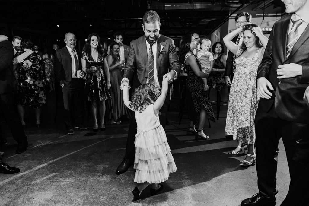 Black and white photo of groom dancing with young girl surrounded by smiling guests during wedding reception at Ovation Chicago
