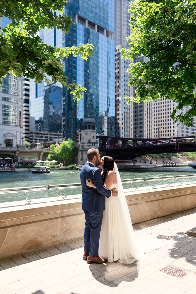 Bride and groom kiss on the Chicago Riverwalk, with city skyscrapers and a bridge in the background