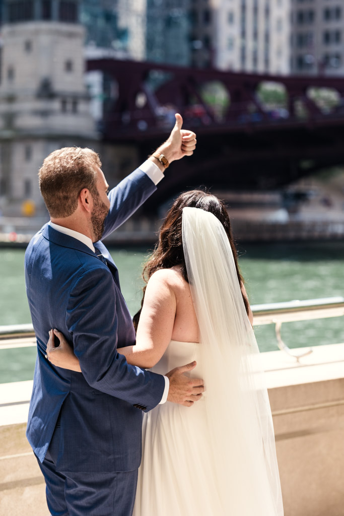 Bride and groom facing away, standing on the Chicago Riverwalk, with the groom giving a thumbs-up to unseen well-wishers