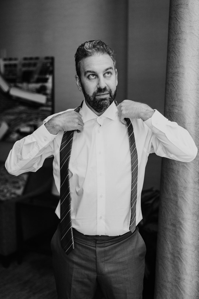 Black and white photo of groom putting on his tie while getting ready for his Ovation Chicago wedding celebration at the Westin River North in downtown Chicago