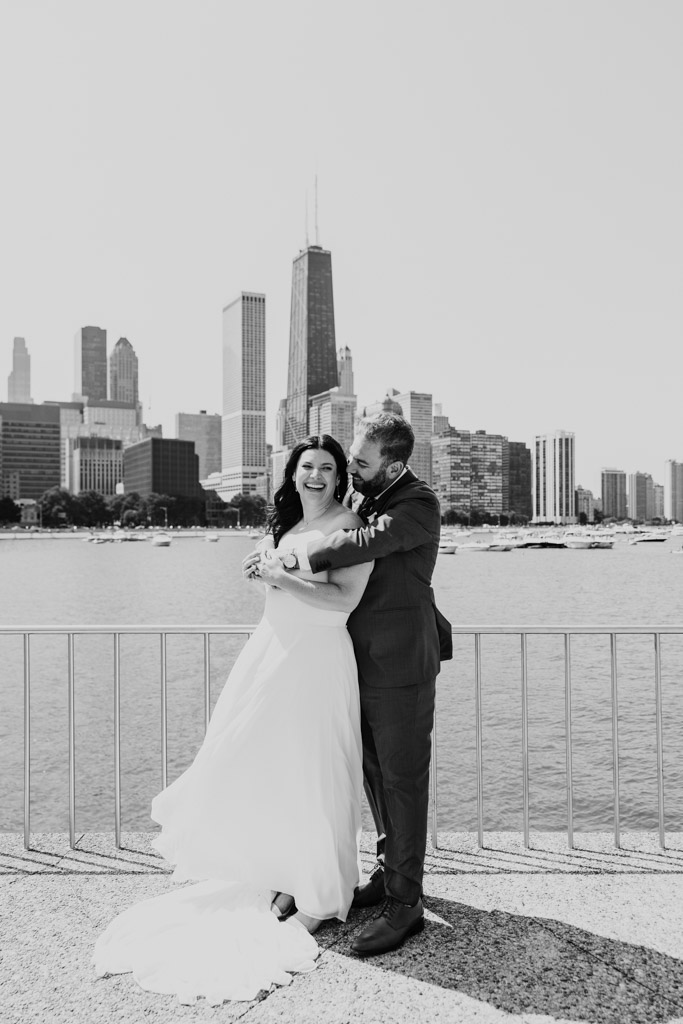 Black and white photo of bride and groom laughing and embracing on the lakefront with Chicago skyline in the background