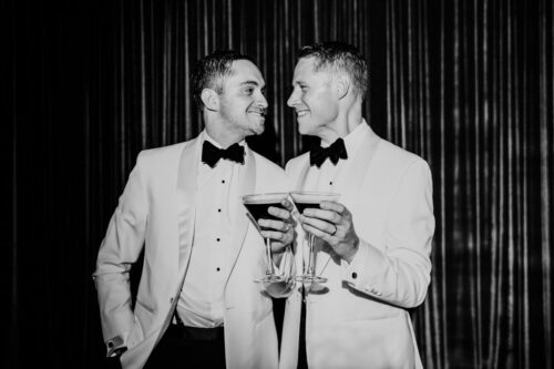 Two grooms in white tuxedos smiling at each other holding espresso martinis on their Chicago wedding day