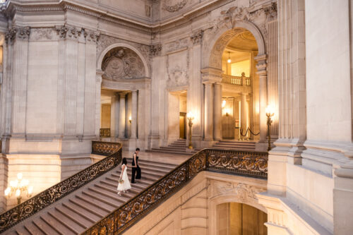 A couple ascends an elegant, grand staircase in San Francisco City Hall with detailed architecture and warm lighting
