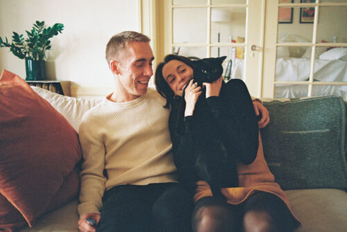 A couple sits on a sofa with their black cat at home during their San Francisco engagement session