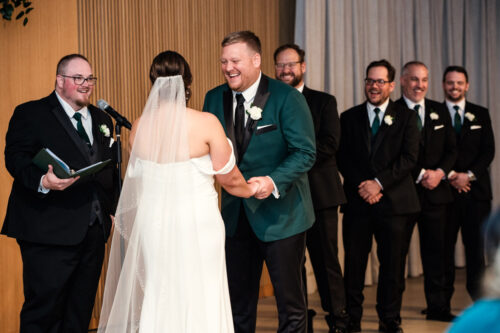 Candid photo of groom in green suit laughing during wedding ceremony at Sarabande Chicago with groomsmen smiling in background