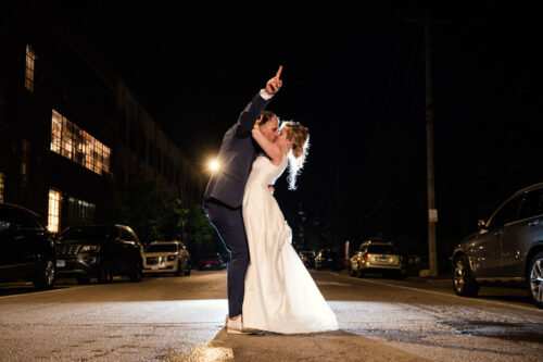 Bride and groom kiss in the middle of a dark street with the groom pointing upward outside The Hive on Hubbard in Chicago wedding reception