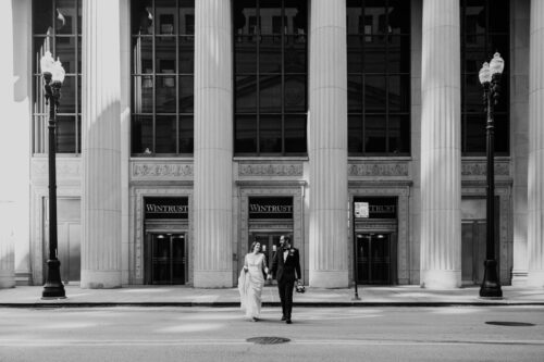Black and white photo of bride and groom crossing LaSalle Street in downtown Chicago hand in hand with a building with tall columns in the background