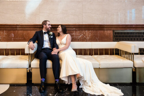 Bride and groom in formal attire sit on a bench, smiling at each other