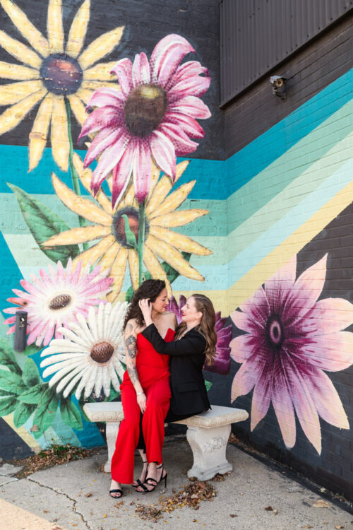 Two people sitting on a bench, one in a red dress, in front of a colorful flower mural.