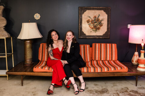 Two women sitting on a vintage striped sofa, smiling, with a dark wall and decorative lamps in the background.