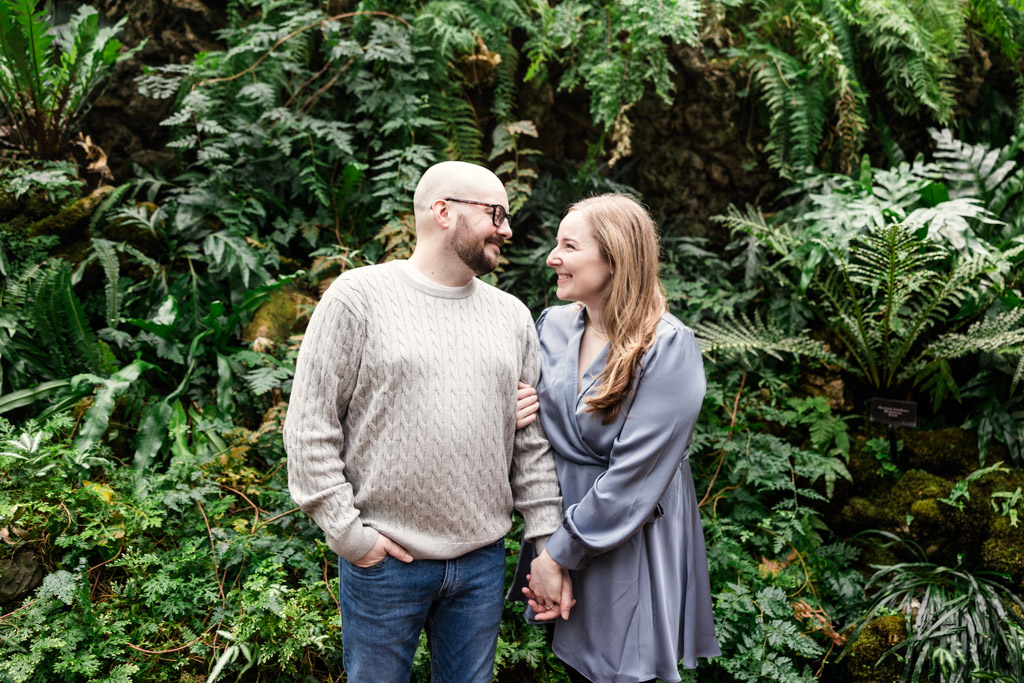 Candid photo of engaged couple standing smiling at each other surrounded by green ferns and plants, inside Lincoln Park Conservatory