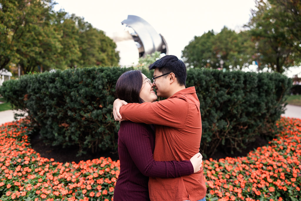 A couple embraces in a garden surrounded by vibrant red flowers, evoking the charm of a Millennium Park engagement, with a sculpture and lush trees gracefully in the background.