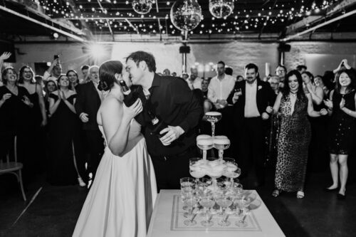 Newlywed couple kisses at their Walden wedding reception, surrounded by cheering guests and a tower of champagne glasses