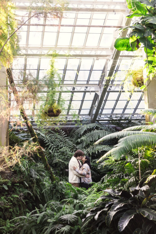 A couple embraces in a lush greenhouse filled with ferns and plants under a glass roof.