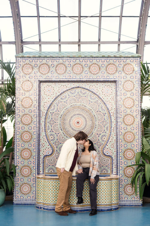 A couple shares a kiss in front of a colorful, ornate mosaic fountain surrounded by plants.