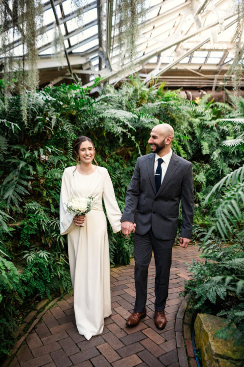 A couple walks hand in hand in a greenhouse, with the woman in a white dress holding flowers and the man in a suit.