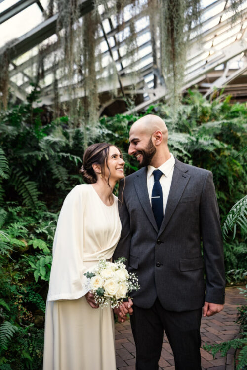 A bride and groom smile at each other in a lush, greenhouse setting. The bride holds a bouquet of white flowers.