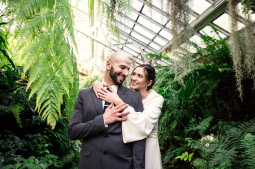 A couple in a greenhouse, smiling and embracing among lush green ferns and plants.
