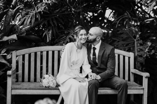 Bride and groom sitting on a bench, groom kissing the bride's forehead, both smiling, surrounded by plants.