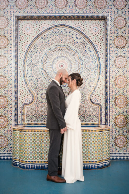 A couple in wedding attire stands holding hands in front of an ornate mosaic wall.