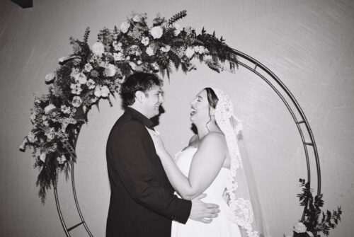 Black and white photo of bride and groom laugh under a floral arch at Walden Chicago after their wedding ceremony