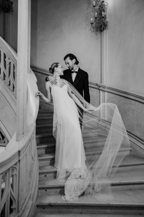 Black and white photo of bride and groom in formal attire pose together on a grand staircase inside Chateau Challain, gazing at each other lovingly during their French castle wedding