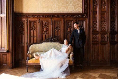 Bride in a white gown lounges on a vintage sofa as a groom in a suit stands beside her in an ornate wood-paneled room inside Chateau Challain