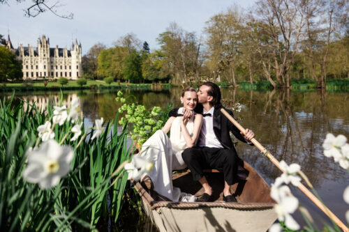 Bride and groom sit in a rowboat on a lake, surrounded by flowers, with Chateau Challain in the background