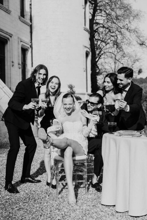 Black and white photo of happy bride and groom posing with four friends outside Chateau Challain, all laughing and holding drinks in celebration