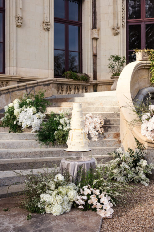 Tall white wedding cake on a table, surrounded by white flowers, near stone steps at Chateau Challain in the Loire Valley, France