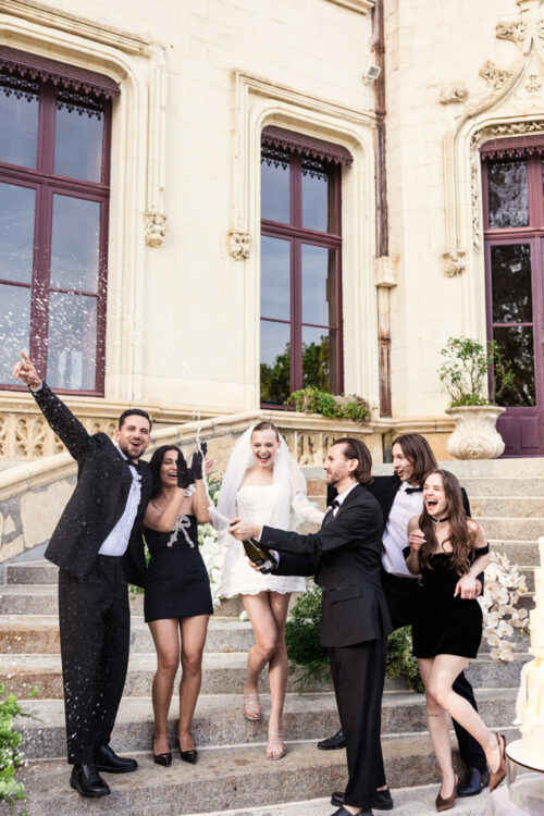 Groom pops a bottle of champagne while bride and the rest of the wedding party celebrate on the steps of Chateau Challain