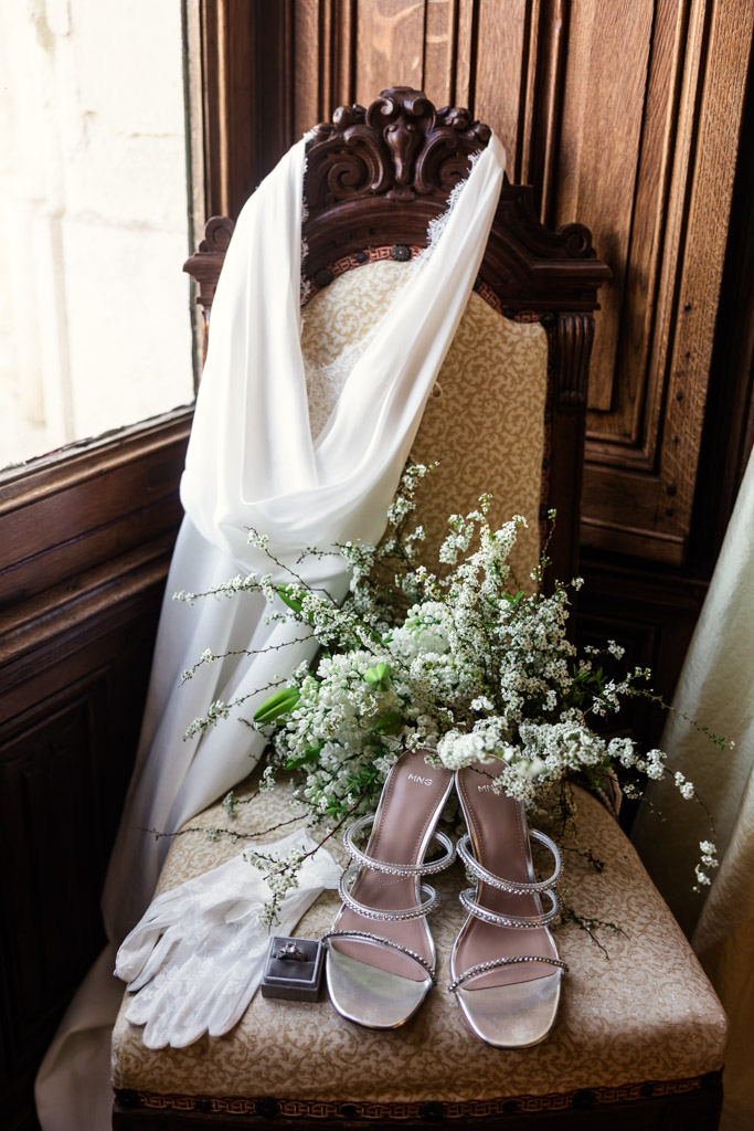 Bride's shoes, gloves, bouquet, and white scarf arranged on an chair by a window inside Chateau Challain