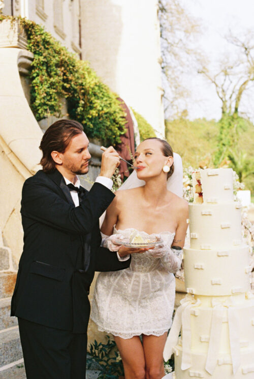 A groom feeds cake to his smiling bride, who holds a plate beside a tall white wedding cake outdoors at their Chateau Challain wedding